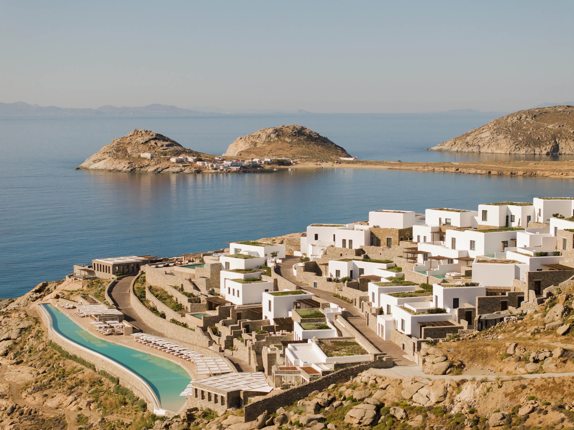 A breathtaking distant shot capturing the entirety of Cali Resort, set against the stunning backdrop of Mykonos’ picturesque landscape, highlighting the resort's luxurious accommodations and the island's natural charm.