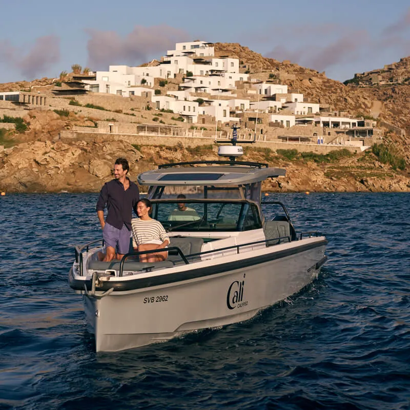 A couple enjoys a scenic boat ride one of Cali's Axopar yachts, cruising away the iconic whitewashed architecture of Cali Resort on the rugged coastline of Mykonos. The setting sun casts a warm glow, highlighting the serene beauty of the Mediterranean setting.
