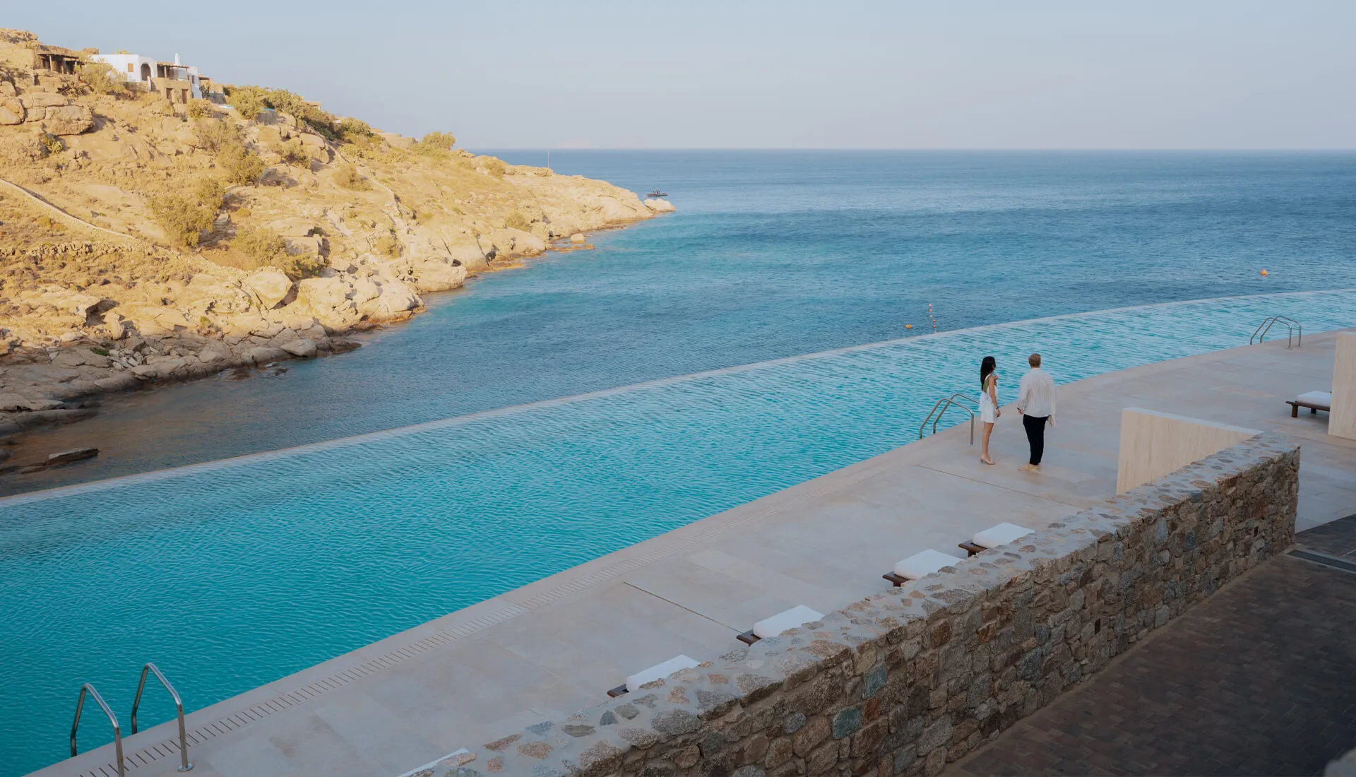 Couple strolling beside the infinity pool at Cali Mykonos, gazing over the serene Aegean Sea during a tranquil early evening sunset.
